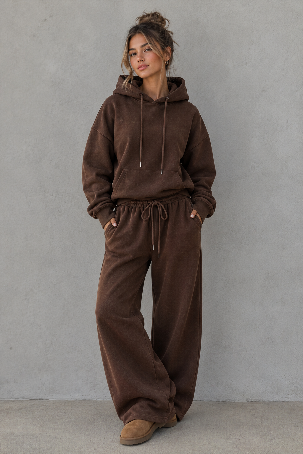 A woman modeling a matching brown lounge set consisting of a pullover hoodie and wide-leg sweatpants, standing against a textured gray wall.
