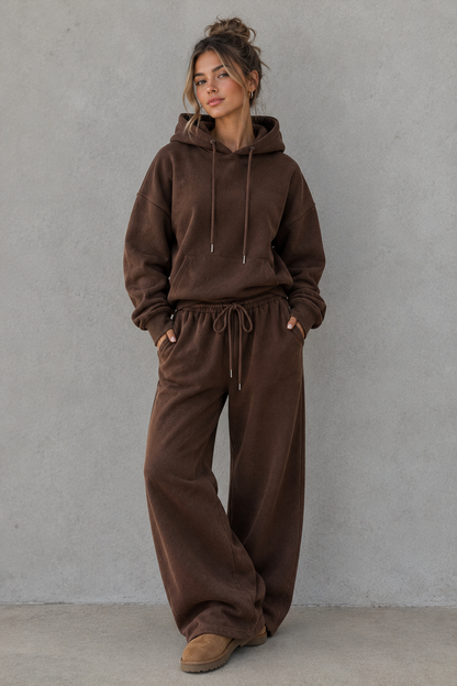 A woman modeling a matching brown lounge set consisting of a pullover hoodie and wide-leg sweatpants, standing against a textured gray wall.